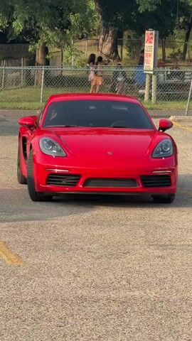 A red Porsche sports car is parked in a lot. A chain-link fence runs along the back of the lot. Beyond the fence, a few people are visible near trees. A sign is posted on a pole behind the car. The car is facing the camera, and the front end is in focus.