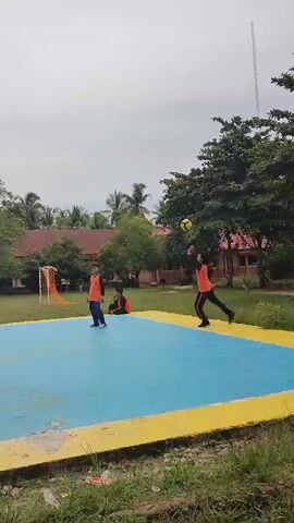 This is a vertical video of people playing volleyball outdoors. The court is blue and yellow, with a net in the center. Several people are on the court, wearing orange shirts, and playing volleyball. In the background, there are trees and buildings. There are also spectators sitting on the grass, some wearing headscarves. The sky is overcast.