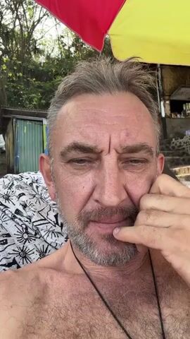 A close-up, selfie-style video shows a man with graying hair and a beard. He is outdoors, with a red and yellow umbrella visible behind him, and lush greenery and a rustic building in the background. The man is looking directly at the camera, touching his face with his hand. He smiles and makes a peace sign with his fingers at one point. The video appears to be shot on a sunny day.
