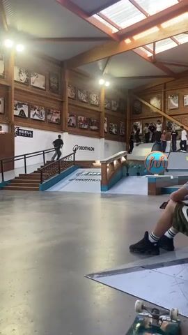 A skateboarder in black clothes does a trick on a metal rail at an indoor skate park. He starts at the top of a small ramp and slides along the rail before jumping off onto a larger flat platform. He then does another trick, jumping off a smaller ramp to land on a box with a metal edge, and then jumping off the other side. The skate park has wood-paneled walls with framed pictures, bright lights, and a skylight roof. A group of people stands on a platform, watching the skateboarder and cheering. There is skate equipment on the ground and in the background.
