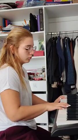 A young woman with blonde hair pulled back and wearing glasses is playing a digital piano. She is wearing a white t-shirt and dark pants. The piano is positioned in front of a closet with clothes hanging inside. The closet is filled with various items on shelves above the hanging clothes, including books and boxes. The scene appears to be indoors, possibly in a bedroom or a living area. The lighting suggests it could be daytime. The woman's expression is focused, indicating concentration on her playing. The overall impression is a quiet moment of musical practice.