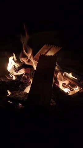 A time-lapse video shows a campfire burning in the dark. The flames flicker and dance, casting a warm glow on the surrounding logs and embers. The fire appears to be burning steadily, with no signs of being out of control. The background is completely black, indicating that it is nighttime or that the camera is focused solely on the campfire. There are no people or animals visible in the frame. The video is very short, lasting only five seconds.