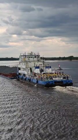 The video shows a pusher tug, marked "OT-2410," with a large flat-top barge in tow, traveling on a waterway under an overcast sky. The barge is loaded with a dark material. The tug is predominantly white and blue. In the distance, a small red and white navigational marker is visible in the water. The water's surface is textured with ripples. Trees are visible in the background along the shoreline.