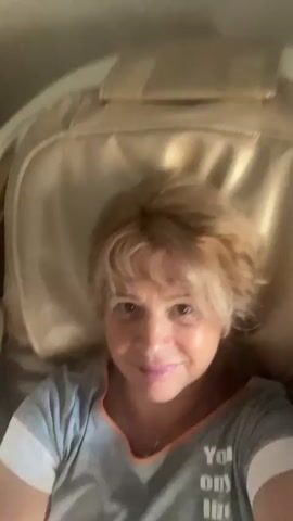 A woman is lying down in a massage chair. She is wearing a gray t-shirt. The massage chair is a light beige color. The video is shaky and out of focus at times. The woman smiles at the camera. The video pans down to show her legs and feet, which are resting in the footrest of the massage chair. Her toenails are painted red. The environment appears to be indoors, possibly a living room or bedroom.