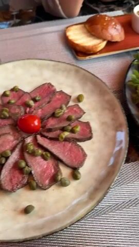 A close-up shot of a table with food and drinks, possibly in a restaurant. In the foreground, a glass of white wine is partially visible, with condensation on the glass. To the left, a plate of thinly sliced roast beef topped with capers is seen. Next to the beef, a plate overflows with fresh strawberries and cherries. In the background, a rectangular platter holds various items, including what appears to be cheese sticks, colorful gummy candies, nuts, and two small dipping bowls, one containing a dark liquid and the other a lighter, paste-like substance. Two hamburger buns are also on the platter. Another person's arm and shoulder are visible in the upper left corner, suggesting someone is seated at the table. The table is covered with a patterned placemat. In the far background, a bouquet of yellow flowers in a vase is visible. The lighting suggests it is daytime, possibly indoors with natural light.