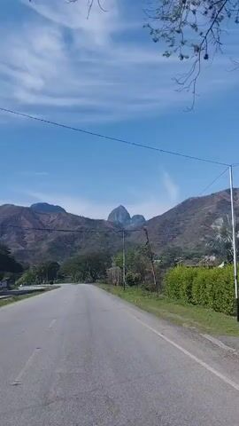 The video shows a drive along a paved road under a bright, clear blue sky with sparse, wispy white clouds. Power lines stretch across the frame, connected to utility poles on the side of the road.

On the right side of the road, there's a vibrant green hedge, followed by a distinctive silver-leaf palm tree. Beyond these, the landscape is dominated by brown and green mountains. Two prominent, cone-shaped peaks are visible in the distance, rising above the other mountains. Some areas of the mountains appear dry or sparsely vegetated.

As the camera moves, a distant settlement or group of buildings can be seen nestled in a valley between the mountains. The road itself appears well-maintained, curving gently through the landscape. The overall scene suggests a rural or semi-rural setting in a warm, sunny climate. No people or visible text are present. The sound of wind accompanies the visual.