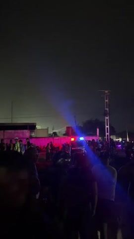 A nighttime gathering of people is visible. The scene is illuminated by colorful lights, including red and blue, which appear to be from police or emergency vehicles. Silhouettes of individuals are prominent, with some faces discernible. In the background, several multi-story buildings are visible, indicating an urban or densely populated area. The ground appears to be uneven and dark. The atmosphere suggests a public event or a situation that has drawn a crowd.