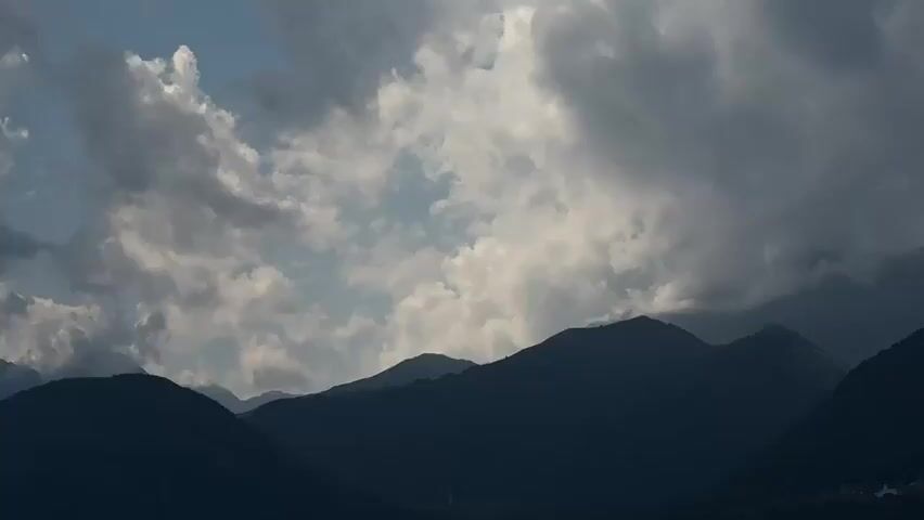 The video captures a sweeping panorama of silhouetted mountain ranges against a backdrop of a dynamic sky filled with dramatic clouds. The foreground features the dark, imposing shapes of mountains, their contours sharp against the lighter sky. In some sections, patches of blue sky peek through the clouds, offering glimpses of sunlight. The cloud formations are varied, ranging from dense, dark masses that cast shadows to lighter, wispy clouds that drift across the scene. The overall impression is one of natural grandeur, with the interplay of light and shadow highlighting the rugged topography of the mountains. There are no visible people or man-made objects in the immediate foreground, emphasizing the natural landscape. The camera pans slowly across the scene, allowing the viewer to take in the vastness of the mountainous terrain and the ever-changing cloudscape.