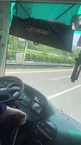 The video shows the inside of a bus from the driver’s perspective. The driver, a young Asian man wearing a blue shirt and black pants, is operating the vehicle on a highway. The dashboard features a steering wheel with the Higer logo, a digital display, and various controls. A Higer-branded sunshade hangs above the windshield. The bus is traveling on a highway with trees and a barrier visible on the side.