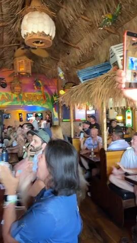 The video shows the interior of a crowded bar with a thatched roof. The bar is decorated with tropical-themed murals and artwork, including a toucan. Several people are seated at the bar and at tables, some of whom are clapping. A television screen in the background displays a sports game or event. A ceiling fan is visible above the bar. The lighting suggests daytime. The atmosphere appears festive.
