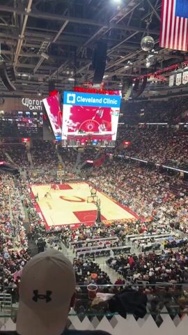 From an elevated vantage point, the video shows a packed Rocket Mortgage FieldHouse in Cleveland during a basketball game. The Cleveland Cavaliers' home court, marked with a stylized "C," is filled with players in action. A massive screen overhead displays the game and advertising for Cleveland Clinic. The stands are teeming with spectators, predominantly wearing casual clothing in various colors. In the foreground, the back of a spectator's head is visible, wearing a white Under Armour baseball cap. An American flag is hung from the rafters.