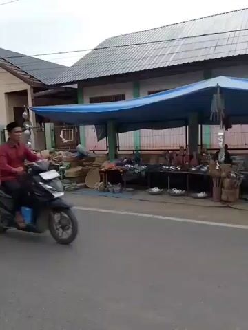 A motorcycle is seen driving past buildings. In the background, there are more motorcycles, people walking, and what appear to be construction materials. The sky is cloudy.