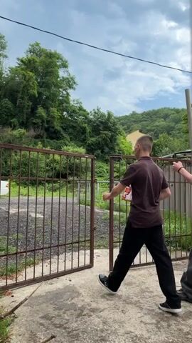 This video shows three young men interacting with a rusty metal gate, likely in a rural or semi-rural setting. The man in the foreground, wearing a brown polo shirt and black pants, is pushing the gate open. Behind him, two other young men are approaching the gate, one in a white short-sleeved shirt and black pants, and another in a white t-shirt and grey pants. The man in the white shirt appears to be holding some documents or a thin book. The setting is outdoors, with lush green trees and hills visible in the background. A yellow building is also visible on one of the hills. The sky is cloudy, suggesting it might be a partly cloudy day. The ground appears to be a mix of concrete and gravel. The overall impression is one of casual interaction, possibly entering or leaving a property. There are no discernible emotions conveyed by the subjects, and no visible text in the frames. The timestamps indicate the video lasts for about 4 seconds. The location context is Shepsi, Russia.