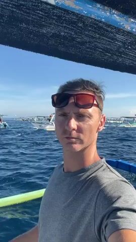 A man in sunglasses and a grey t-shirt is standing on a boat. He looks directly at the camera. In the background, several outrigger boats bob on a calm, blue sea. The sky is clear. Above the man's head, a dark mesh provides shade. The man lowers his right hand from a wave.
