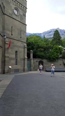 The video captures a courtyard with cobblestone paving. On the left, a portion of a stone building with a round clock face is visible. A man in a white t-shirt and khaki shorts walks away from the camera towards the center of the courtyard. Further into the courtyard, a woman in a floral dress and a man in a dark t-shirt and jeans stand together, facing away from the camera, with another woman in a white top and khaki pants standing to their right, holding a phone as if taking a photo. In the background, there is a stone wall topped with lush green trees and foliage. Beyond the greenery, majestic mountains with rocky peaks rise against a cloudy sky. There are several benches along the stone wall. Several other individuals are visible in the distance, scattered throughout the courtyard, also appearing to be tourists. The overall scene suggests a historical or scenic tourist destination.