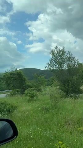 The video shows a drive through a green landscape on a partly cloudy day. The view from inside the car shows a winding road ahead and rolling hills in the distance. Green grass and trees line the road, and the sky is filled with a mix of white and dark clouds against a blue background. A side mirror is visible in the lower left corner of the frame.