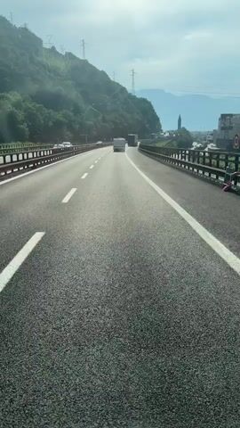 The video shows a view from a vehicle traveling on a highway. The highway has multiple lanes and is bordered by guardrails. On the left side of the highway, there is a steep, tree-covered hillside with power lines running along it. On the right side of the highway, there are buildings and more guardrails. In the distance, mountains are visible under a cloudy sky. Several other vehicles, including trucks and cars, are visible on the highway ahead. The perspective suggests the video is recorded from the driver's or passenger's seat of a vehicle.