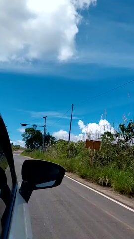 The video shows a car driving along a winding road under a bright blue sky with scattered white clouds. The camera is positioned inside the car, showing the side mirror. The road is asphalt with a white line marking the edge.  Vegetation, including tall grass and bushes, lines the side of the road. Utility poles and power lines are visible. A sign is present next to the road, but the details are obscured. A motorcycle can be seen in the distance ahead on the road. The sound of wind is prominent.
