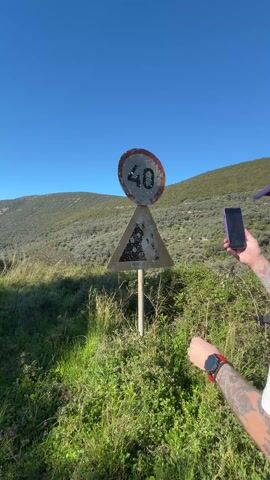 A man with a backpack is photographing a bullet-ridden road sign with a smartphone. The man is wearing a white t-shirt with a graphic design on the back, a backwards baseball cap, and has tattoos on his arm. He is holding a purple smartphone and taking a picture of the triangular sign, which has a round speed limit sign above it, both covered in bullet holes. He reaches out and touches the sign with his hand. The background features a hilly landscape with green vegetation and a clear blue sky.
