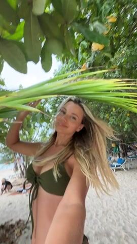 The video shows a blonde woman in a green bikini on a sandy beach. She is holding a large palm frond over her head and smiling at the camera. In the background, there are lounge chairs and other beachgoers. The scene is tropical, with lush green trees and foliage surrounding the woman.