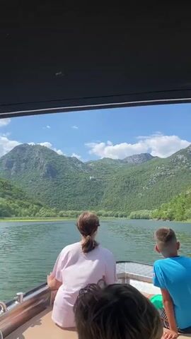 The video shows a view from a boat on a body of water. In the foreground, the backs of two people are visible, a woman with her hair in a ponytail wearing a light pink shirt, and a young boy in a blue shirt. The boat's railing is also visible. The background features a wide expanse of water leading to a lush green mountainside. A white building with a red roof is visible high on the mountain, alongside a road with parked cars. The sky is blue with scattered white clouds. The camera appears to be moving slowly forward, with the boat creating ripples on the water. The overall impression is a scenic boat tour on a sunny day.