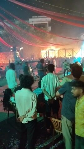 The video shows a film set at night. A crowd of people, some of whom are crew members holding cameras, stand watching a performance on a brightly lit stage. The stage is set against a background of scaffolding and artificial fog. Red fabric is draped above the crowd.