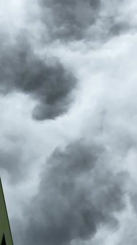 The video shows a low-angle shot of a gray, cloudy sky with dark, amorphous smoke or clouds billowing and shifting. On the left side of the frame, the corner of a light green building with dark rectangular windows is visible. The camera is static, focusing on the sky and the building's facade. No people or distinct actions are observable beyond the movement of the atmospheric elements.