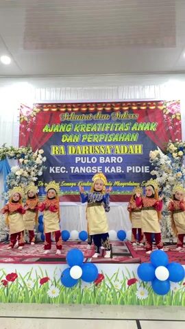 This video shows a group of young children performing a dance on a stage. The children are dressed in traditional attire, including gold crowns and intricately designed clothing in red and gold. One child stands in the center, performing a solo dance, while the others are arranged around them, participating in the group dance. The stage is decorated with blue and white balloons, and a large banner in the background reads "SELAMAT dan SUKSES AJANG KREATIFITAS ANAK DAN PERPISAHAN RA DARUSSA'ADAH PULO BARO KEC. TANGSE KAB. PIDIE". This translates to "Congratulations and Success Children's Creativity Event and Graduation RA Darussa'adah Pulo Baro Tangse Sub-district, Pidie Regency". The event appears to be a graduation or end-of-year celebration for a kindergarten or early childhood education center. The setting is indoors, likely a hall or auditorium, with white walls and ceiling lights visible. The atmosphere seems festive and celebratory, with the children appearing engaged and focused on their performance. The backdrop suggests a formal event with a significant banner indicating the occasion and the institution involved.