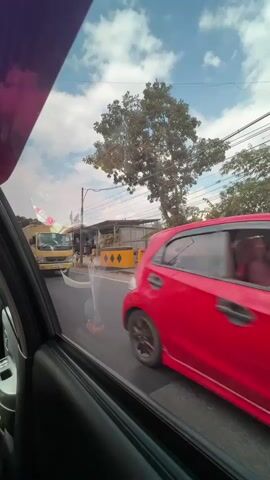 The video shows a yellow truck with a shipping container traveling on a road. The camera is inside a vehicle, looking out the window. A red car is also visible in the foreground, moving in the same direction. The road appears to be in an urban or suburban area, with trees and buildings in the background. The sky is blue with some clouds. The truck is the primary focus of the video, and its movement and details are captured as it passes by.