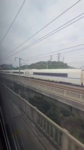 The video shows a view from a moving train. In the foreground, a dark, blurry barrier runs horizontally across the bottom of the frame. In the middle ground, a long, white high-speed train with blue accents travels on an elevated track. The train has multiple cars and appears to be in motion. Beyond the train, a lush green hillside is visible under a cloudy sky. Electrical wires crisscross the upper portion of the frame, suggesting the train is operating on an electrified line. The perspective is from a window, indicating the viewer is inside another train.