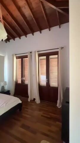 The video pans across a bedroom with a slanted, wood-paneled ceiling. The room features a bed with white linens, two bedside tables with lamps, and a dark headboard. On the wall above the bed is a framed piece of art. Two sets of French doors with wooden blinds are visible, covered by white curtains. The floor is made of polished wood. To the left, a white roller blind covers a window. Further left, a tall, wooden wardrobe with silver handles is seen. A doorway leads to another room, revealing a toilet and sink. The video also briefly shows a white object hanging from the ceiling near the center of the room.