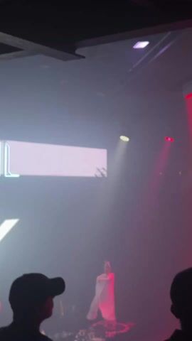 A dark, smoky nightclub scene unfolds.  The focus is on a figure, likely a performer, illuminated on stage by both a bright white spotlight and red stage lighting. The performer is silhouetted against the light, and they appear to be wearing white attire with what could be wings or flowing fabric.  In the foreground, silhouetted heads of audience members can be seen. A man wearing a baseball cap is visible in the center of the frame.  The club's branding, featuring a neon "L" and other illuminated elements, is partially visible on the left. There are moments of brighter red light shooting up from the stage toward the ceiling. The audio includes music and a voice hyping up the crowd.