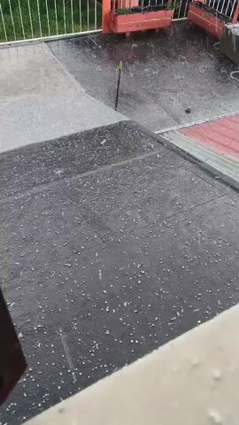 The video shows an outdoor elevated area, likely a balcony or rooftop terrace, during a daytime hail storm. In the foreground, a dark gray/black flat roof or balcony surface is covered in numerous small white pellets of hail that are actively falling and bouncing. Further back, a lighter gray concrete-like surface is also visible and appears wet. On the far side of the roof area, two red wooden planter boxes with green plants can be seen. A white metal railing runs along the edge of this section, with green foliage visible beyond it in the background. A green stick-like object stands upright near the planter boxes. To the right, red corrugated metal roofing is partially visible. The surfaces are wet and reflective due to the precipitation. The primary activity is the ongoing hail storm, with the sound of hail hitting the surfaces audible. No people or visible text are present. Given the context, this scene is set in Quito, Ecuador.
