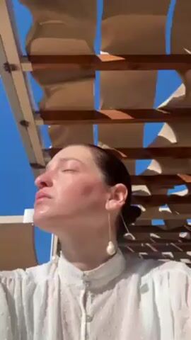 A woman is looking up, her face in the sunlight. She has fair skin, and her hair is pulled back. She is wearing a white, semi-sheer blouse with a high collar, and she has a dangling pearl earring. She is standing under a pergola-like structure with a slatted wooden frame and fabric panels that provide some shade. The sky is blue.