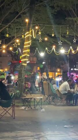 A group of people are gathered outdoors at night, illuminated by string lights and star-shaped decorations hanging from trees. In the center, two women are standing, one of whom is singing into a microphone. Several other individuals are seated in folding chairs, facing the performers. Behind them, a colorful inflatable structure and a stage area are visible. The overall atmosphere suggests an outdoor performance or gathering.