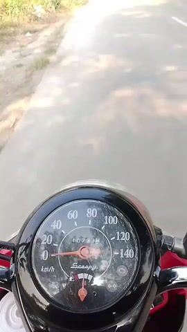A first-person perspective video of someone riding a scooter on a paved road lined with trees. The scooter's speedometer is visible, showing a speed of approximately 20 km/h. The road appears to be a rural or suburban area, with vegetation on the sides. The sky is bright, suggesting a sunny day. There are other scooters or motorcycles visible further down the road. The rider's shadow is cast on the road ahead.