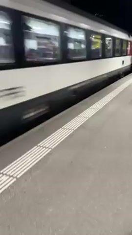 The video shows a train moving along a railway platform. The train is white with a red stripe. The train is labeled SBB CFF FFS. The platform is gray and has white textured stripes. The video is taken from a low angle. The video is taken at night.