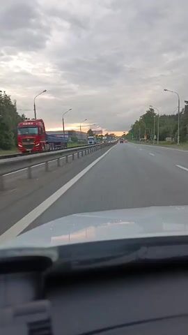 This video captures a drive along a highway in Monino, Russia. The perspective is from inside a vehicle, looking out through the windshield. The time of day appears to be late afternoon or early evening, with a cloudy sky tinged with the warm colors of a setting sun visible on the horizon. The road is a multi-lane highway with guardrails separating the lanes and the opposing traffic.

On the left side of the highway, there are trees, industrial buildings, and streetlights. Several large trucks are also visible on the highway and on the side of the road, suggesting a commercial or industrial area. On the right side, there are trees and some road signage, including what appears to be a speed limit sign. The activity is simply driving on the highway, with other vehicles visible in the distance, indicating moderate traffic.

The weather is overcast, but the light suggests a break in the clouds or the sun is low enough to cast a glow. The overall mood is neutral, capturing the mundane experience of highway travel. There is no discernible text that is clear or legible enough to be read.