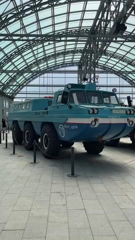 The video shows a large, amphibious vehicle displayed indoors under a geodesic dome structure. The vehicle is teal and white with six large, rugged tires. It has a cabin with windows at the front and a long, flat body. Several stanchions with ropes are placed around the vehicle. The floor is tiled with light gray square tiles. In the background, other parts of vehicles are visible, as well as the metal framework of the dome. The lighting is bright and appears to be natural light coming from the dome. There are no people clearly visible interacting with the vehicle. The overall impression is that of a museum or exhibition setting.