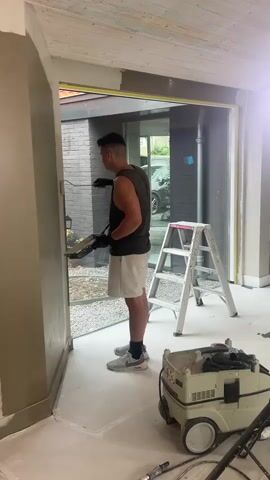 A man is standing on a white floor, wearing a dark sleeveless shirt, white shorts, and sneakers. He is holding a trowel and a small container, seemingly applying a substance to a wall. He appears to be working on an interior renovation project. To his right, a silver step ladder stands tall, and behind him, a large window offers a view of an outdoor area with gravel and a dark brick building. A beige wall frames the left side of the image. In the foreground, a beige and white industrial vacuum cleaner with wheels is parked on the floor, with some cables connected to it. The ceiling above is made of light-colored wooden planks. The scene suggests a construction or renovation site, possibly in a residential setting. The lighting appears to be natural, indicating it might be daytime. There is no visible text in the media file. The overall mood is one of focused work.