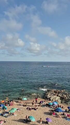 The video shows a crowded beach on a sunny day with a clear blue sky and scattered white clouds. The turquoise water is calm with small waves breaking near the shore. A rocky outcropping extends into the sea on the right side of the frame. Many people are sunbathing on the sand, some under colorful umbrellas. Several individuals are in the water, swimming or wading. In the distance, a boat with a parasailer is visible on the ocean. The beach is covered in light brown sand and has a variety of beach umbrellas and towels.