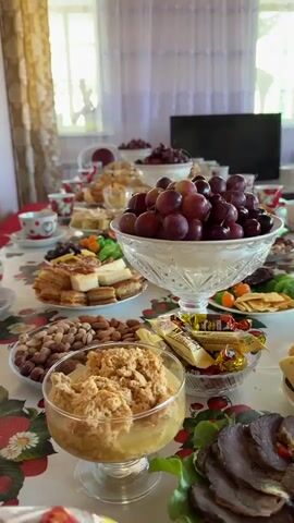 A white tablecloth with a red fruit design is seen covering a table laden with an array of food. In the foreground, a glass bowl holds a brown dessert, and a plate of sliced meat is visible. The camera slowly pans to the right, revealing more dishes. A large glass bowl filled with red grapes is prominently displayed in the center. Surrounding it are plates of various pastries, nuts, dried fruits, and candies. In the background, a window with sheer white curtains is visible, and beyond it, some greenery can be seen. A dark television screen is positioned in the far background. The table is set with numerous teacups and saucers, suggesting a gathering. The room has light-colored walls.