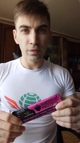 A man in a white t-shirt holds a pink packet of "SUCCESS PROTEIN" in his left hand while flexing his right bicep. The packet features a graphic of a strawberry and text in a foreign language. The man is looking directly at the camera with a slight smile. The background appears to be an indoor setting with furniture and a window.