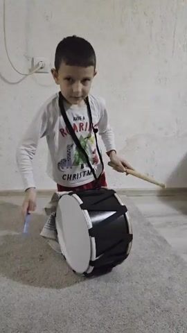 Here's a description of the video:

A young boy is playing a black and white drum. He is kneeling on a gray rug and holding the drum in front of him with a strap around his neck. He is using two drumsticks, one blue and one wooden. The boy is wearing a white long-sleeved shirt with a Christmas themed graphic and red pants. The background consists of an off-white wall with an electrical outlet visible. The floor is tiled.