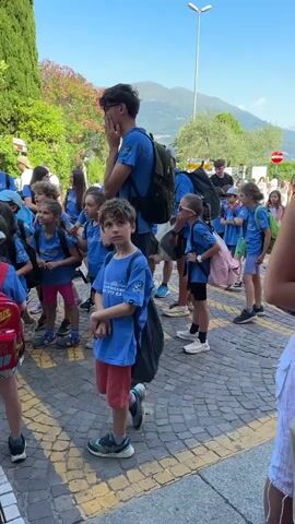 A group of children and young adults in blue t-shirts are standing on a cobblestone street. Most of the children are wearing backpacks. The youngest child in the foreground is wearing red shorts and a blue t-shirt. The tallest young adult in the background, also wearing a blue t-shirt and backpack, is looking up and appears to be speaking. The scene is outdoors, with trees and a mountain visible in the background. The sky is blue and the sun is shining. The children appear to be waiting or listening to instructions.