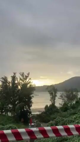 The video shows a cyclist riding a bicycle near a barrier, with a view of a body of water, trees, and a distant cityscape. The scene shifts to a first-person perspective of someone riding a motorcycle on a road with cars parked on the side. The rider wears a helmet and a backpack.