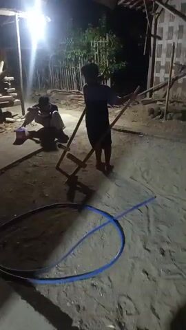 Two young males are in an outdoor, dimly lit area at night. One male, wearing a dark t-shirt and shorts, is standing on a wooden contraption that appears to be stilts. He is holding a long stick or pole vertically. The other male, wearing a lighter colored shirt and pants, is sitting on the ground to the left of the standing male, leaning against the wooden contraption. He is holding a red and blue container. A blue hose is visible on the ground in the foreground. The background shows wooden structures and bamboo poles. The scene appears to be in a rural or undeveloped setting.