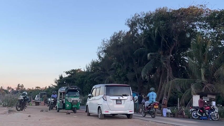 The camera slowly pans from left to right in a seaside town. At the beginning of the video, a white minivan and two green tuk-tuks are visible. In the distance, there are trees and a few people standing and sitting around. As the camera pans further, a black tuk-tuk with the words "Surf Juice Arugam Bay" on the back becomes visible. The camera focuses on a green-and-yellow building called "Elephant House," surrounded by palm trees. Nearby is a small, thatch-roofed structure where people are sitting. Motorcycles are parked in front of both structures. A large green pillar is also visible near the thatched structure. In the background, there are more trees and the sky is clear and blue.