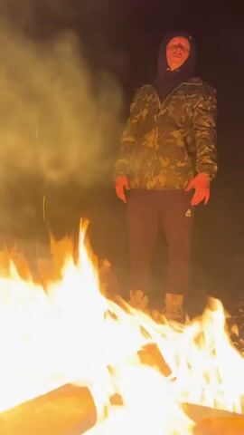 A man stands in front of a large bonfire at night. He wears a camouflage jacket over a hooded sweatshirt, dark pants with a logo near the ankle, and red gloves. The camera is positioned low, looking up at the man, making the fire appear larger. The background is dark, with a faint suggestion of a crescent moon in the upper left. The man is looking towards the camera. He speaks and the camera zooms into the bonfire.