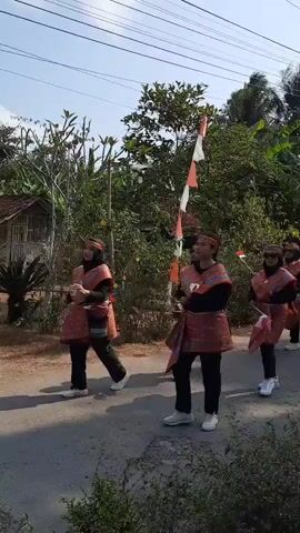 A group of five individuals in matching cultural attire walks down a paved road. The attire consists of black pants, black long-sleeved shirts, red and gold patterned sashes worn diagonally across the chest, and matching red and gold patterned skirts or wraps over the pants. They are also wearing head coverings, some of which appear to be black hijabs, and white sneakers. The individuals are mostly facing to the right of the frame, with some turning their heads to look at the camera. They appear to be marching or parading, with some holding small Indonesian flags. The road is lined with lush green vegetation, including trees and bushes. In the background, there are more trees, a few simple structures, and utility poles with wires overhead. A string of red and white flags, likely Indonesian flags, is strung across the road on a pole in the middle ground. The sky is a pale blue with a few wispy clouds. The overall impression is a cultural procession or celebration in an outdoor, rural setting.